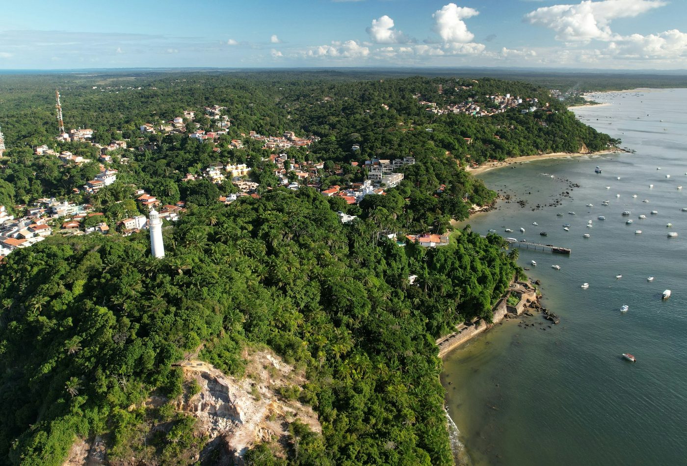 Vista aérea do farol de Morro de São Paulo sobre o morro coberto de mata, com a enseada e barcos ao fundo