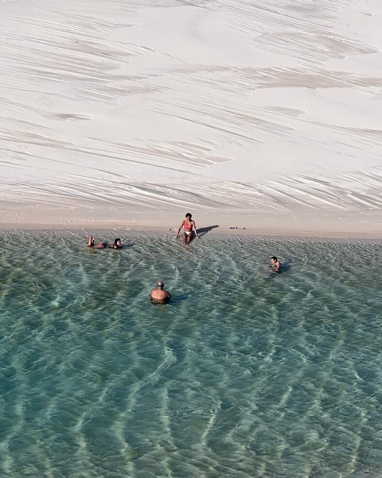 Lagoa rasa e transparente aos pés de uma duna, com banhistas ao longe