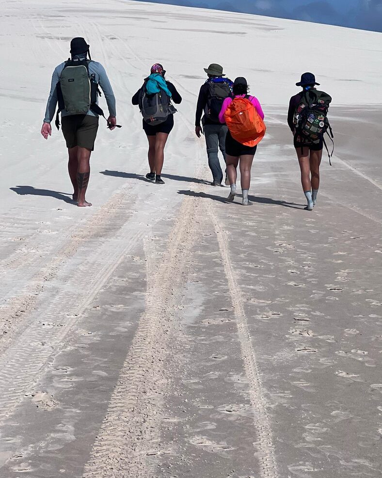 Grupo de visitantes caminhando em fila sobre areia batida rumo às dunas