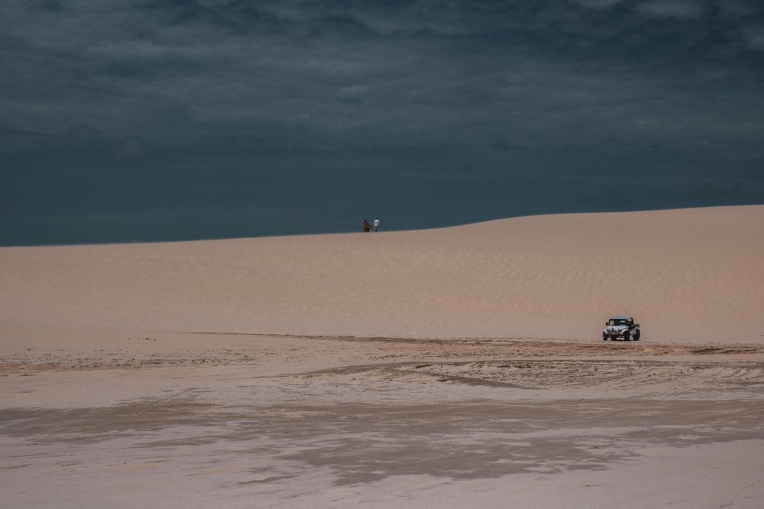 Veículo 4x4 sozinho numa faixa larga de praia molhada, com dunas ao fundo