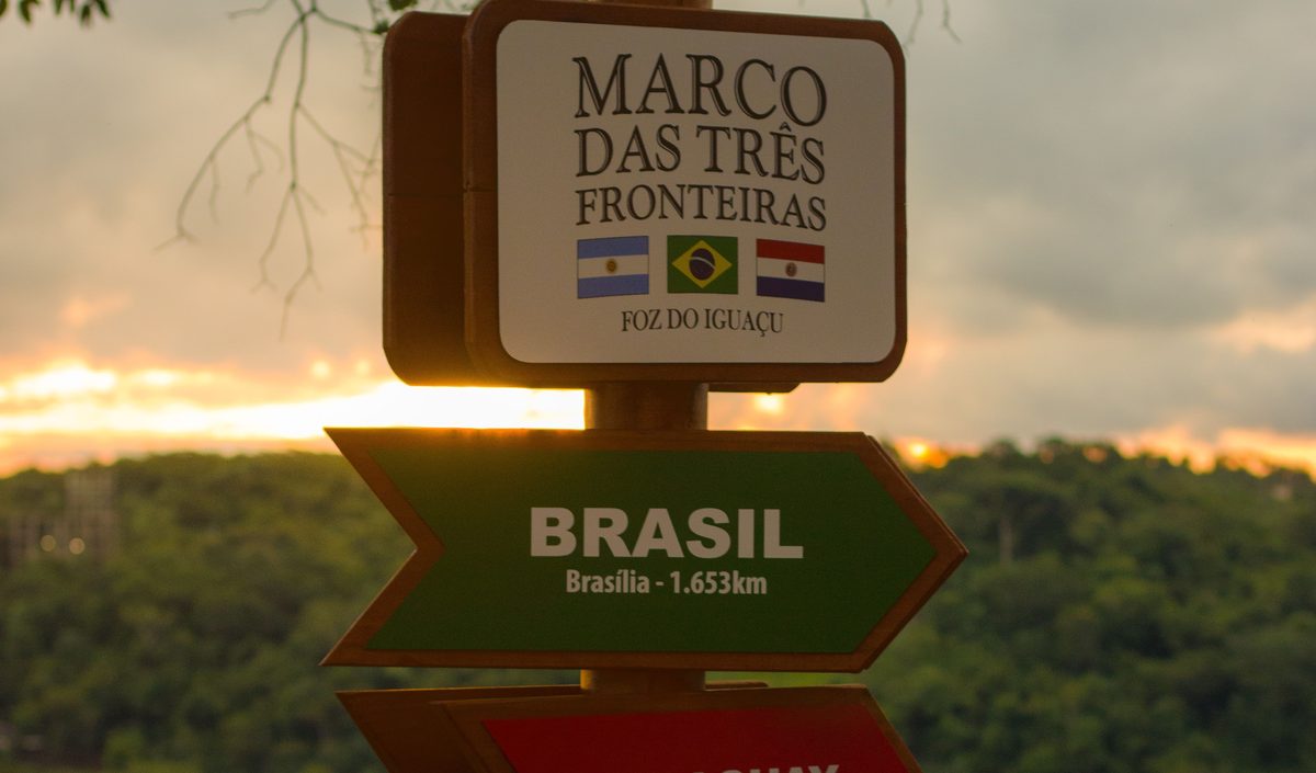 Placa do Marco das Três Fronteiras, em Foz do Iguaçu, com o pôr do sol sobre o rio Paraná ao fundo
